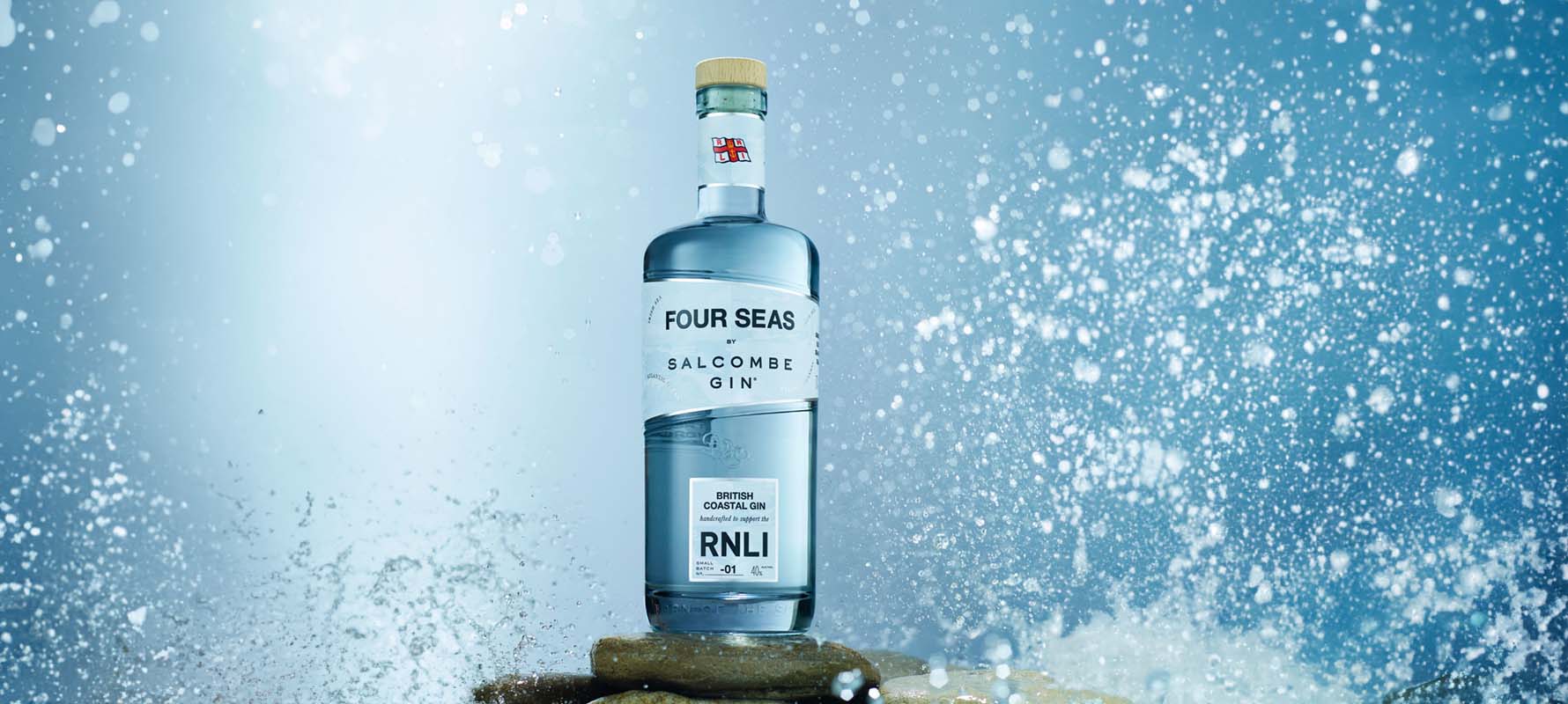 A bottle of ‘Four Seas’ gin by Salcombe Gin, in partnership with the RNLI, is on a rock with sea splashing around it.