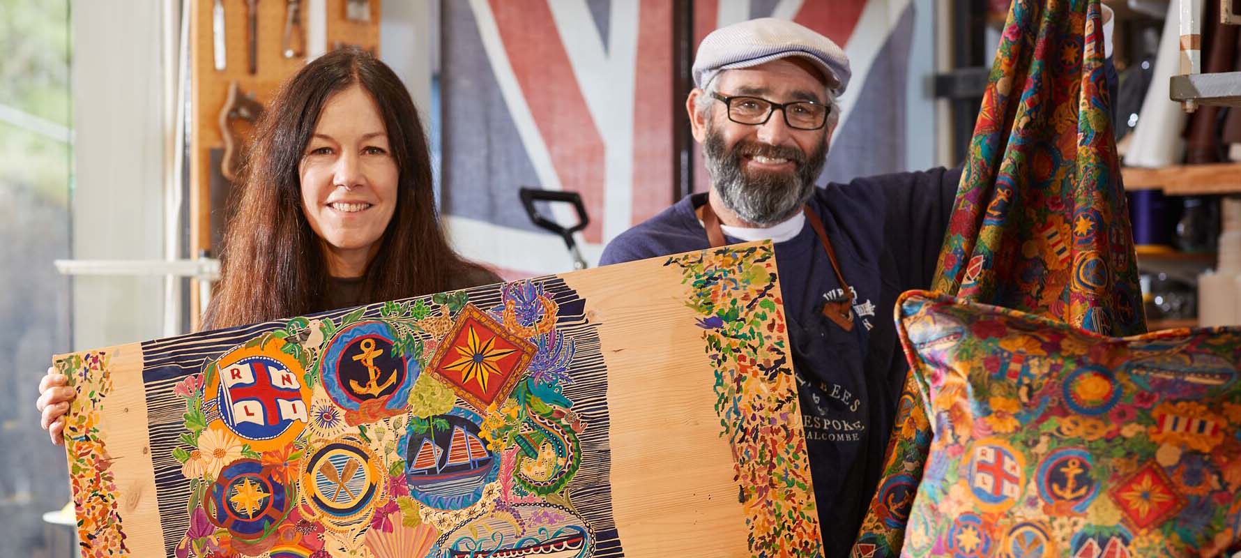 A woman and a man are stood in a workshop and are holding up products with a colourful, intricate design inspired by the RNLI.