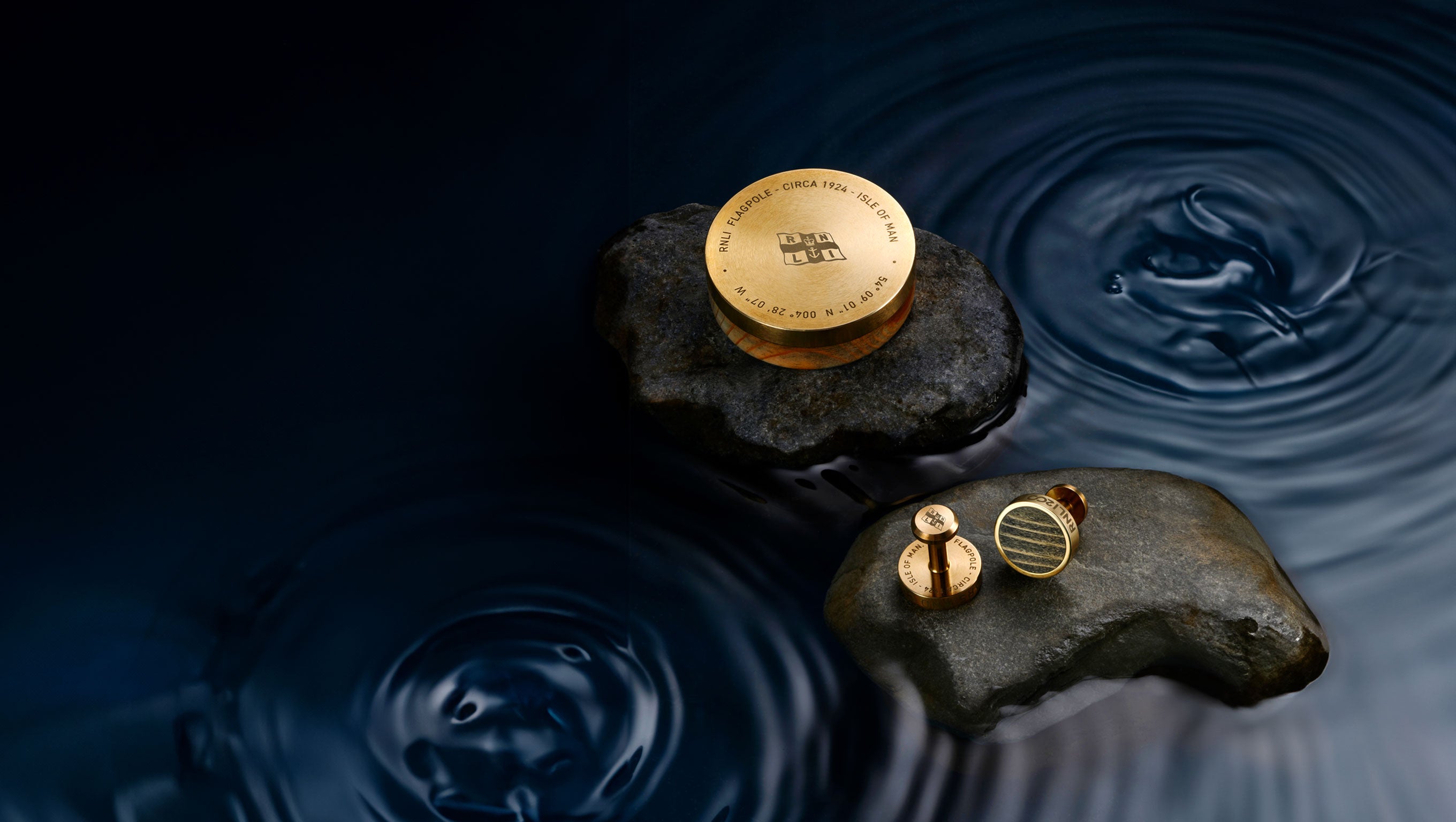 A brass and wooden RNLI branded paperweight and cufflinks are displayed on rocks, with water around them.