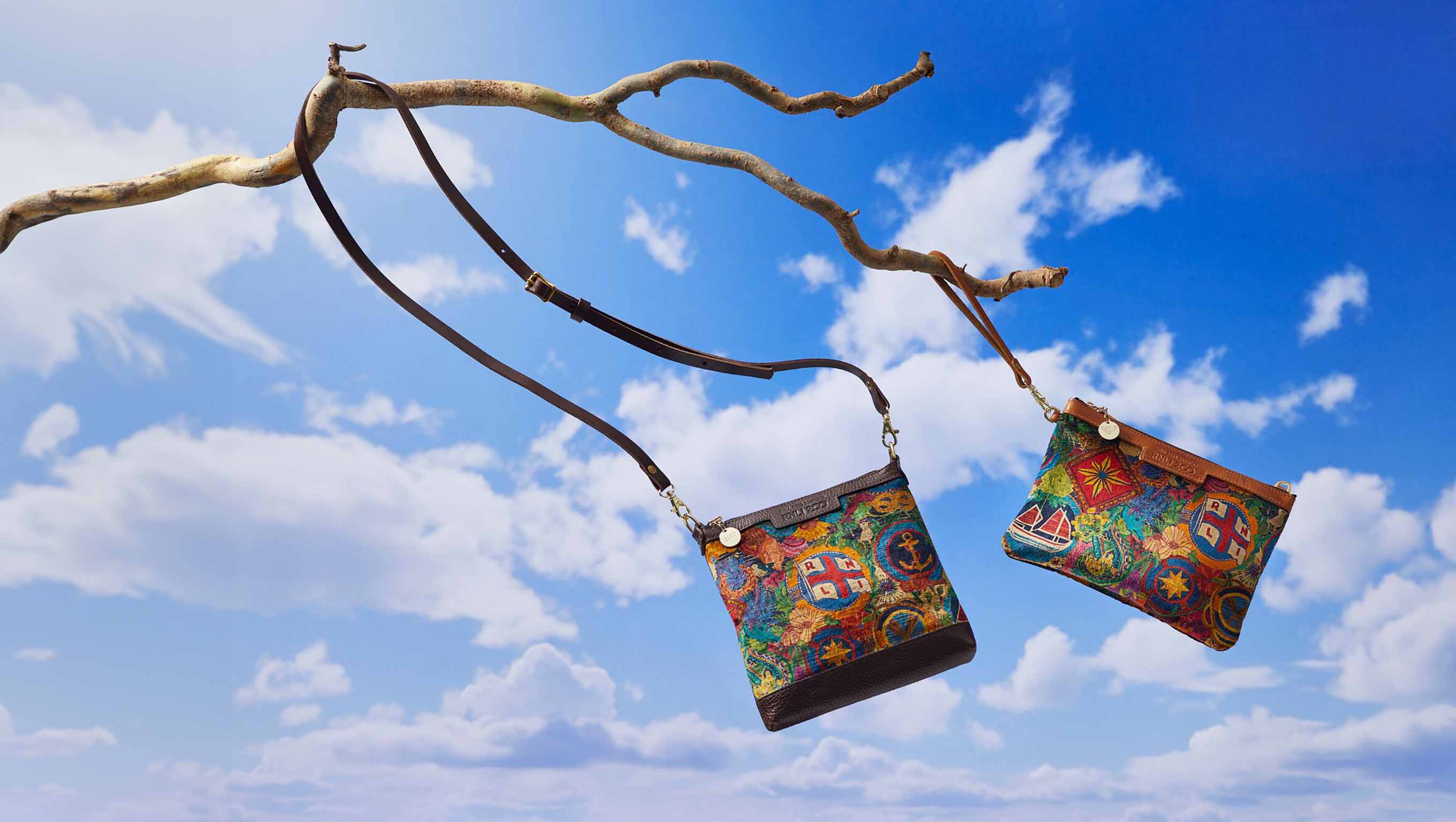Two clutch bags featuring colourful RNLI designs are hanging from a tree branch and swinging in the wind, with blue sky and clouds in the background.