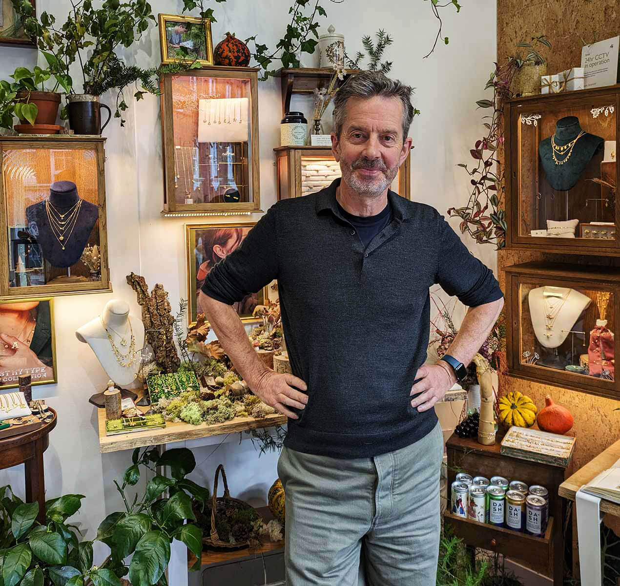 Alex Monroe is stood with his hands on his hips and smiling with jewellery display cabinets and indoor plants around him.