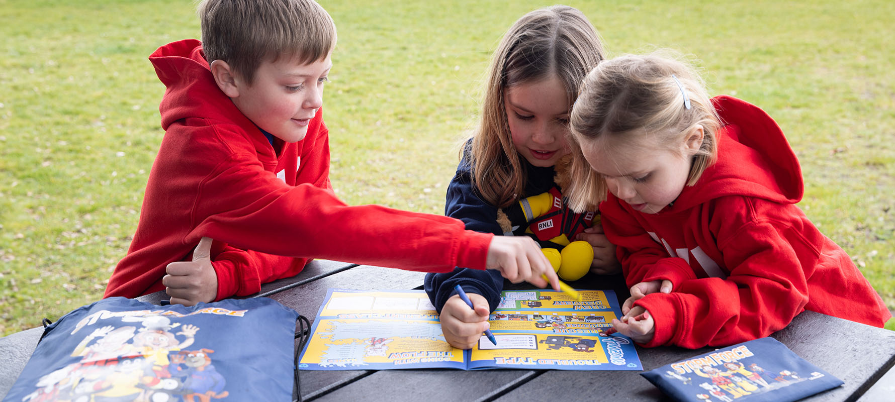 Three children are sitting at a table outside on the grass. They are crowding around a Storm Force magazine, and one of the children is using a pen to write in the magazine. There are beach huts in the background.