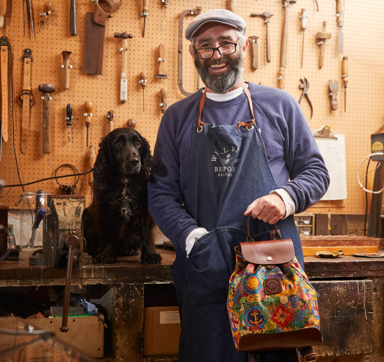 Will Bees is stood in his workshop holding one of the RNLI 200 Limited Range Rucksacks, with his black dog sat on the worktop next to him.