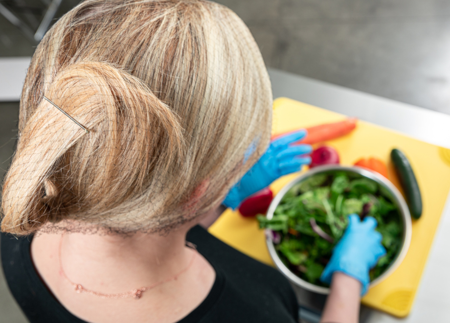 a white person with long blonde hair prepares a salad. they are wearing a black hairnet and blue disposable gloves