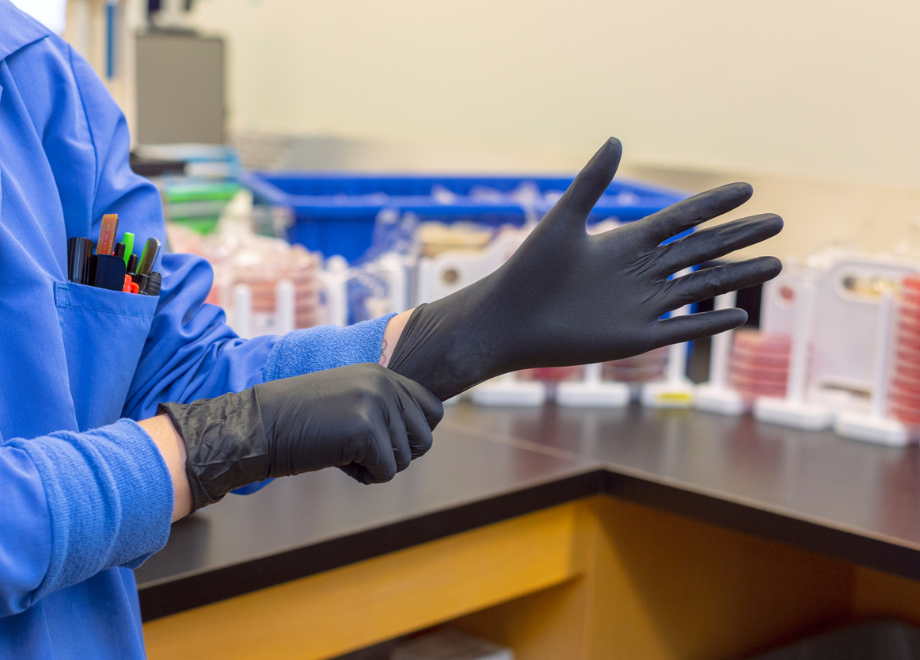 Person wearing a blue lab coat is working in lab. They are putting on a pair of disposable black nitrile gloves. The person is a white adult with tattoos on their arms. 