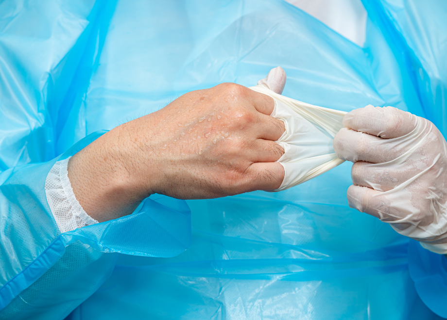 A person wearing an isolation gown and cover is doffing a glove from their right hand. Their hands are visibly sweating