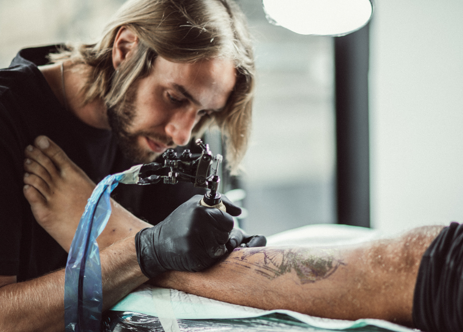 A man with long hair and a beard is safely tattooing another person's left shin. We can't see the rest of the person being tattooed