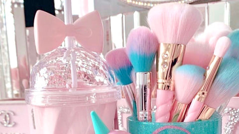 Belle bow cup third image shows the blush pink cup with transparent spill proof cover and signature bowtie on a desk, next to teal and pink makeup brushes in a sparkly coffee mug. Bright light and background mirror emphasize the glitter aesthetic.
