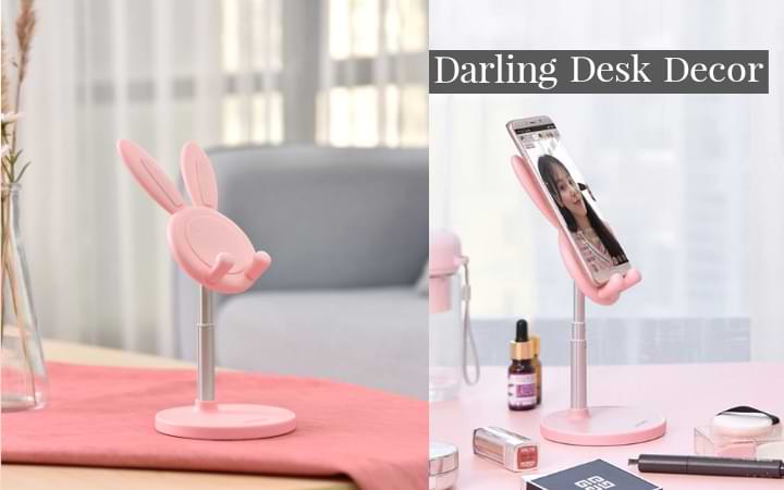 Two images of the pink Boston Bunny Phone Stand on different desks: the stand on a pink textured fabric with grey background furniture, then the stand holding a phone on a white countertop with other pink makeup accessories surrounding it
