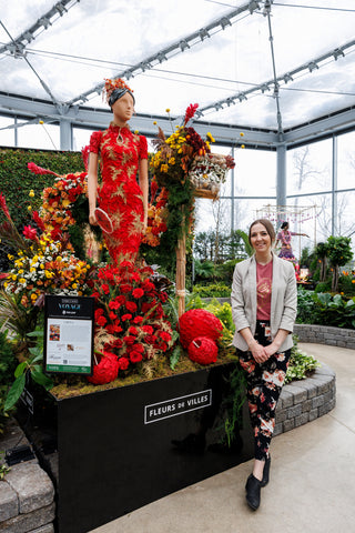 woman standing in greenhouse space with mannequin in chinese dress of red rose petals with gold leaf detail.  Dancing Dragon made of flowers  wrapped around the display.  Plants along a winding path in background.