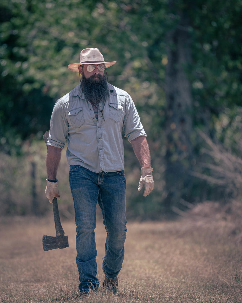 Image of a working man walking while holding an ax. He's in jeans, a long sleeve button-up, gloves, boots, glasses and the Barcelona |  Wide Brim Straw Sun Hat by American Hat Makers