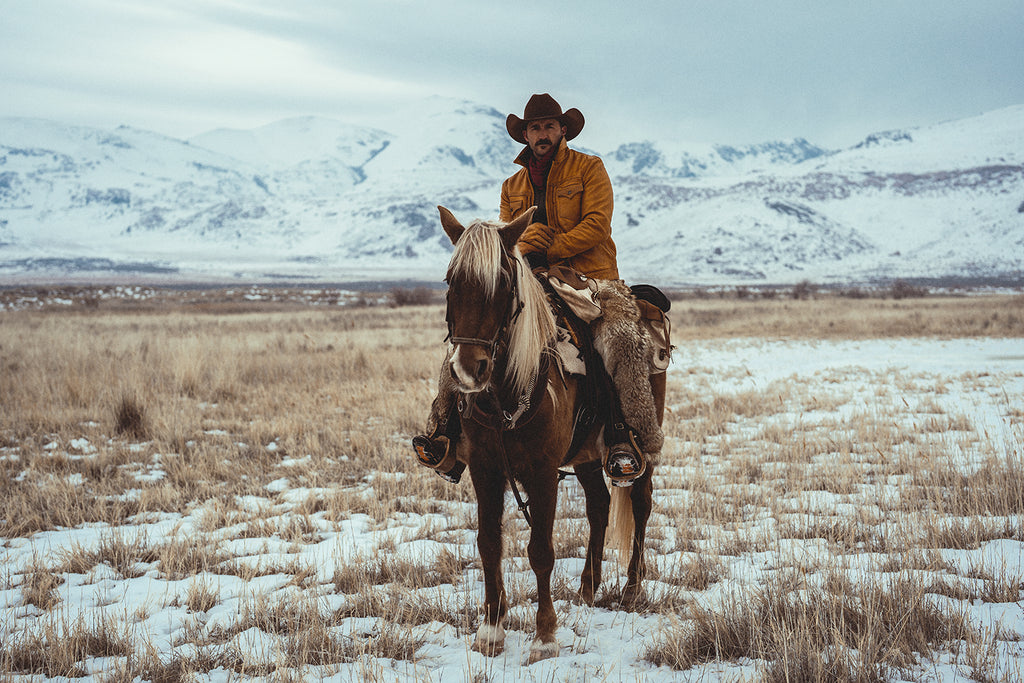 a snow dusted field with a male cowboy on horse who's wearing fur chaps, a mustard colored jacket and a brown felt Cattleman hat by American Hat Makers