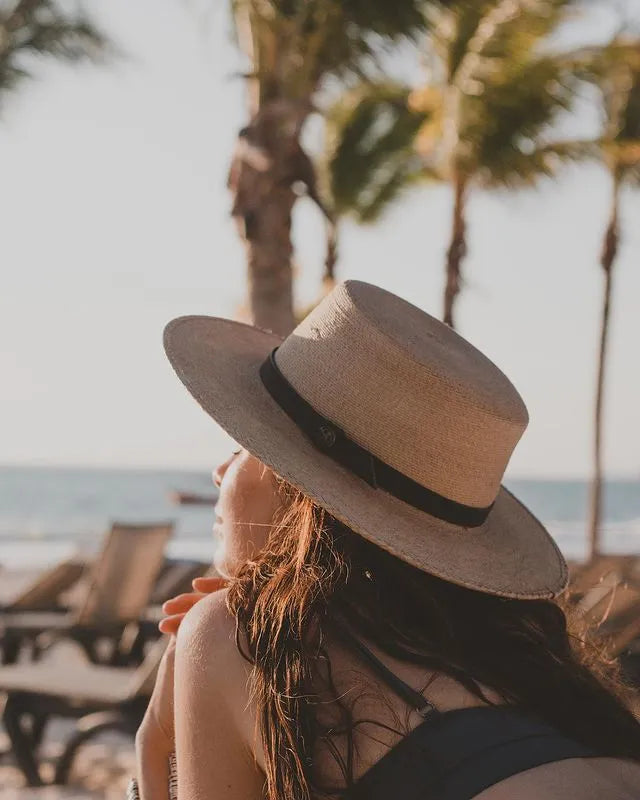 image of girl lying on the down on her stomach in the sand while looking out towards the water wearing a black bikini top and the Cozumel Wide Brim Straw Hat by American Hat Makers