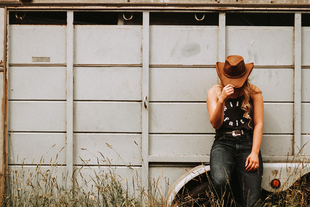 blonde haired girl leaning on horse Trailor with grass as high as her knees in a black tank top that says "wild faith", blue jeans  and her hand tipping down the front of the copper colored Hollywood Cowboy Hat by American Hat Makers