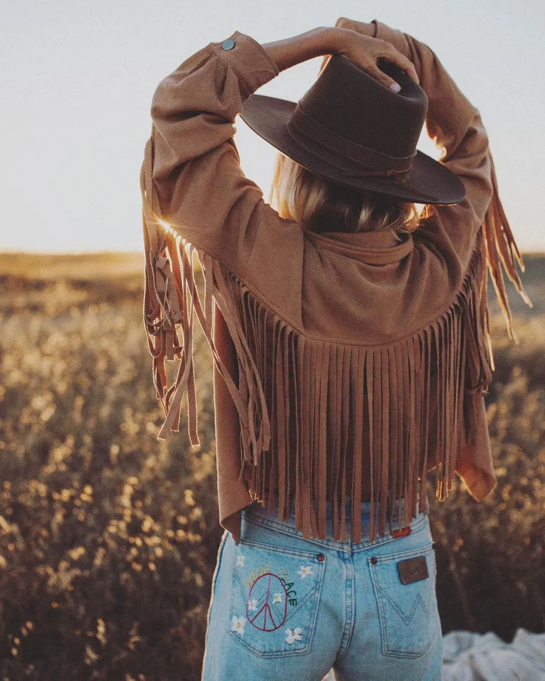image of back of girl wearing a brown fringe jacket, blue jeans and her arms above her head with her hands resting on a brown felt fedora hat from American Hat Makers (Java Hat)