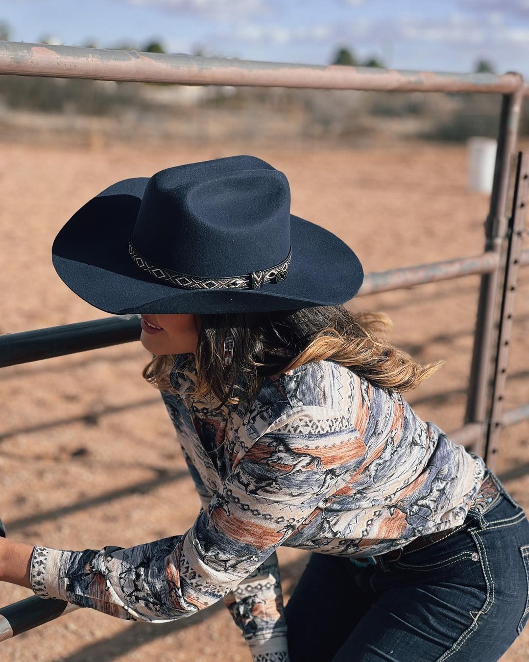 girl with brown hair in a horse print shirt and blue jeans peering through a metal post horse arena wearing the Jessy, a navy felt cowboy hat by American Hat Makers
