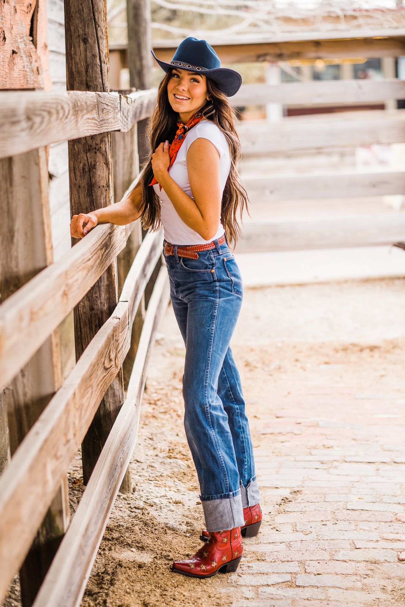 girl with brown hair in a white t-shirt, blue jeans and a red belt, red print scarf and red cowgirl boots with yellow stars wearing the Jessy | a navy felt cowboy hat by American Hat Makers