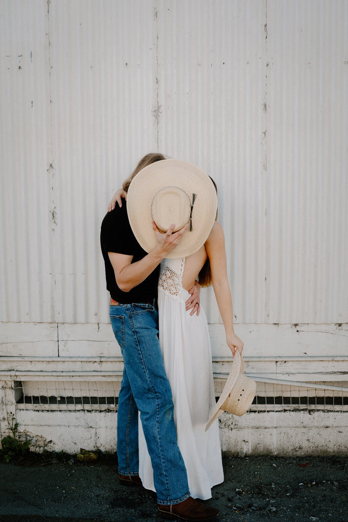 image of a couple kissing, she is wearing a long white dress and he's wearing blue jeans, a black shirt, covering their faces with an Elkhorn, a straw cowboy by American Hat Markers