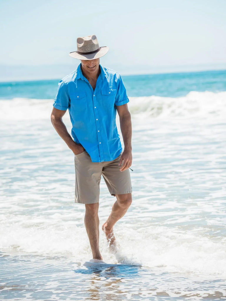 image of guy walking on along the beach with the water just covering his feet, he's wearing khaki colored shorts, a blue button up short sleeved shirt and the Milan | Men's Straw Fedora Hat by American Hat Makers