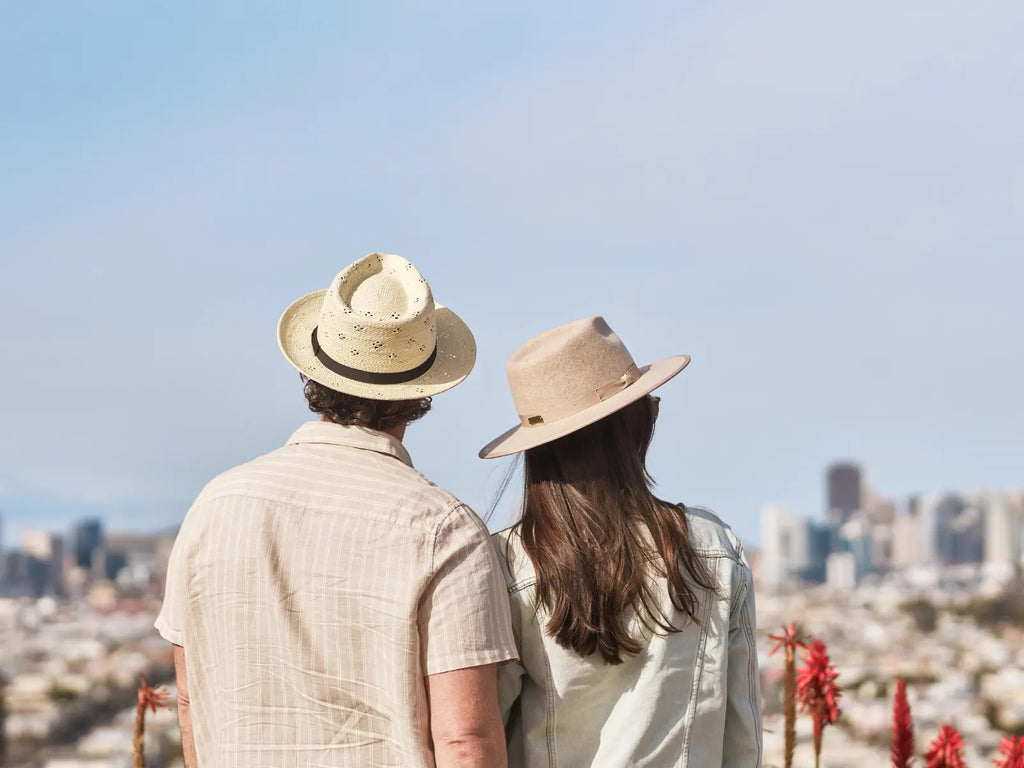 Image of a couple looking over a city. He's wearing a cream button up t-shirt with white stripes and a Dimitri Straw Fedora Hat in Natural color by American Hat Makers. She's wearing a white jean jacket and wearing the Bondi Wide Brim Felt Hat in Oatmeal color by American Hat Makers