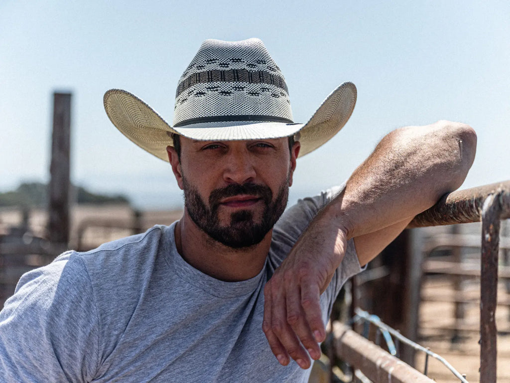 Image of a cowboy resting his arm against a metal fence in a grey t-shirt wearing the Cisco Straw Cattleman Cowboy Hat by American Hat Makers