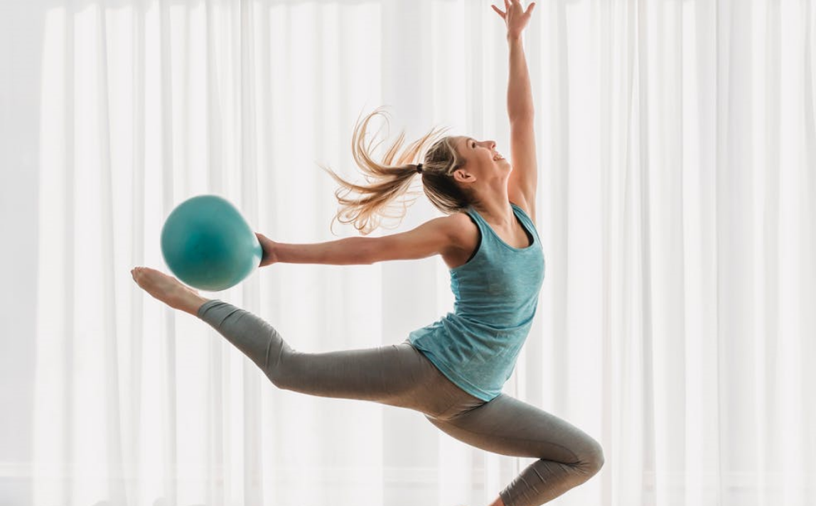 A rhythmic gymnast is in an acrobat position while holding a ball in her right hand