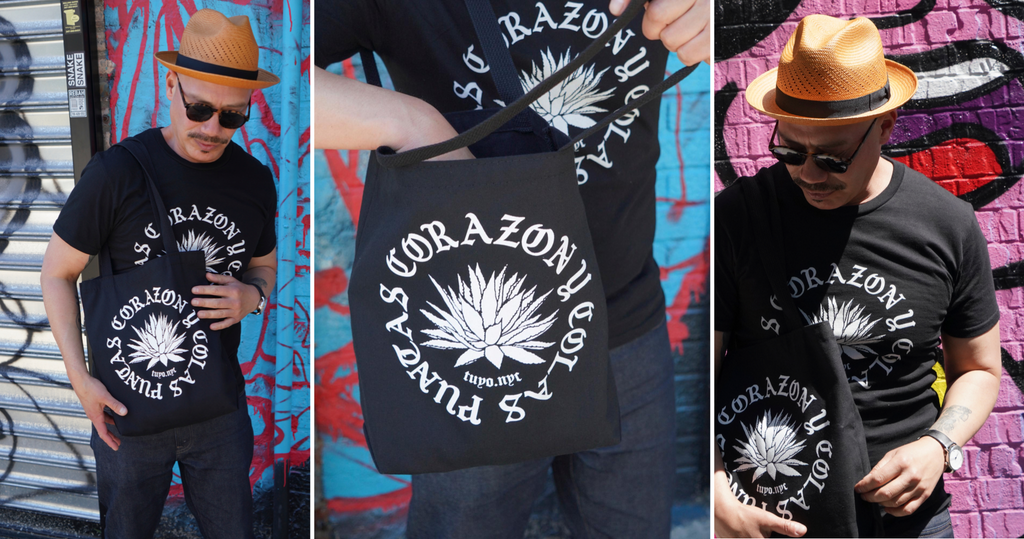 Black cotton t-shirt worn by male model who is holding a black tote bag with the same design. The design is printed in white featuring the text: puntas, corazon and colas in a circle format with an agave plant illustration inside the circle.