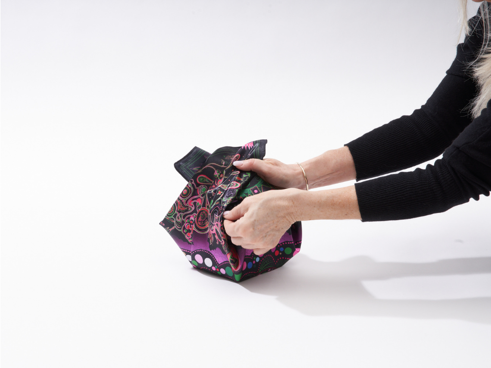 Imagen de alguien preparándose para atar las esquinas opuestas de un envoltorio furoshiki sobre una caja de regalo.
