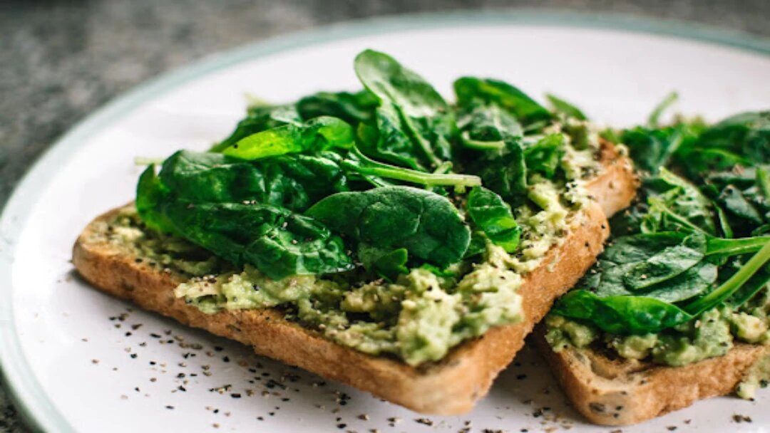 A white plate with toast with avocado and spinach on top.