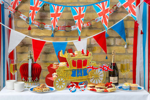 Coronation buffet table ready for a street party with blue paper napkins and plates, royal carriage cake stand and hanging red, white and blue bunting.