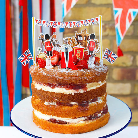 Tall Victoria sponge with Coronation themed cake toppers on including King Charles, Jack Russell's, Royal carriage and Union Jack flags 