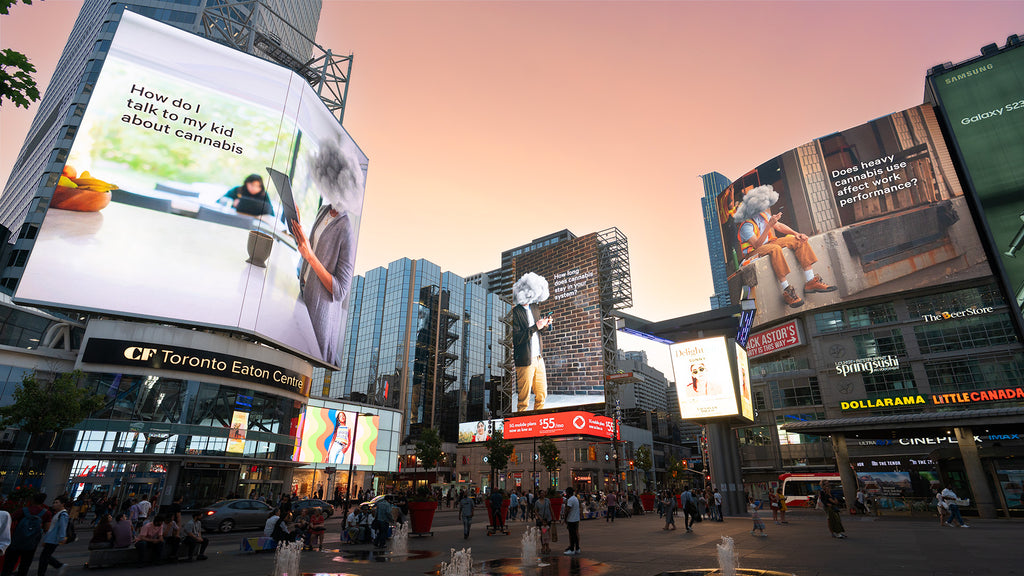 Photo of large, digital billboards featuring Cannabis Made Clear campaign content as they illuminate Yonge and Dundas Square in Toronto. The image is taken at street level where dozens of pedestrians are walking. The sky above the large city buildings is pink at sunset.