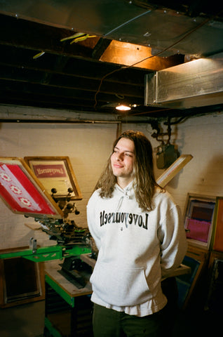 A man wearing a hoodie stands next to a screen printing press