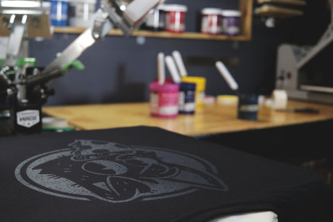 a black shirt printed with white ink sits on a press with colored inks nearby