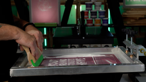 A hand pulls a squeegee on a screen with pink emulsion