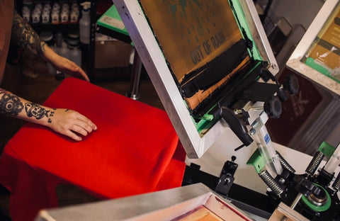 A hand rests on a red shirt loaded onto a screen printing press