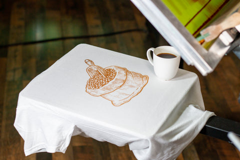 A screen printed bag of coffee grounds on a white T-shirt with a cup of coffee 