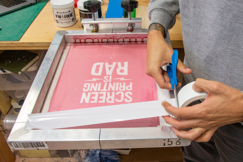 a hand cuts tape off a screen that says "screen printing is rad"
