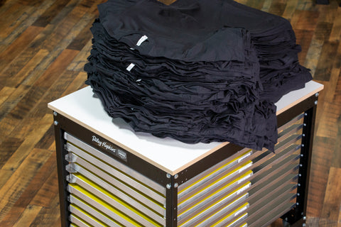 Black t-shirts sit on a press cart