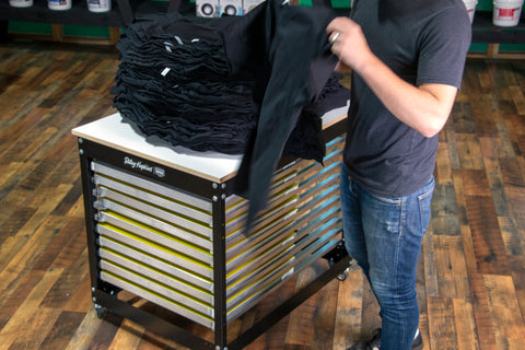 A man holds up a black t-shirt from a pile laying on a press cart