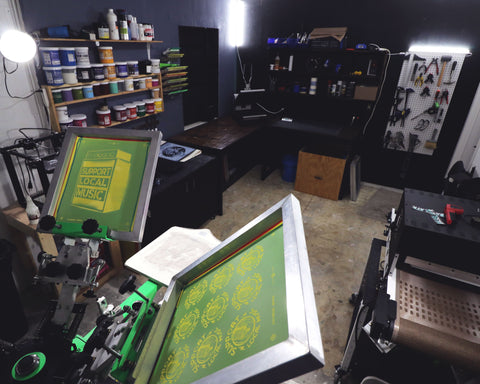 A screen printing press sits at the foreground of the photo, with a conveyor dryer and wall of ink behind it