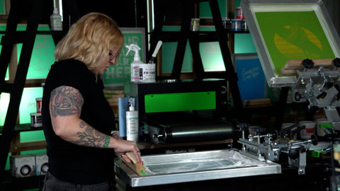 a woman with blond hair pushes a squeegee of white ink across a screen