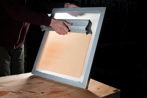 A hand holding a scoop coater and coating a screen with orange emulsion