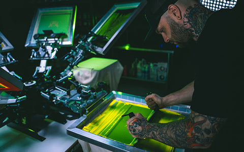 a man wearing a hat with neck tattoos pulls an EZ grip squeegee over a screen on a riley hopkins 250 press