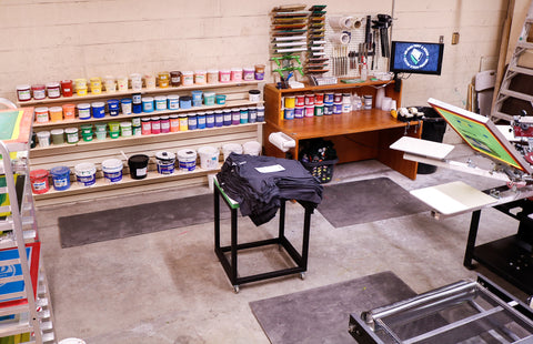 A shop with a press, ink wall, and shirts sitting on a cart