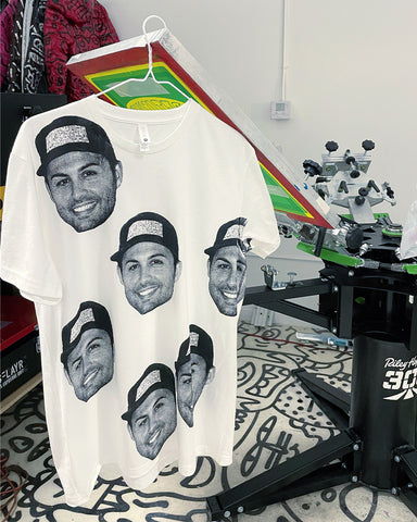 a white t-shirt with faces on it hangs from a screen on a riley hopkins 300 press