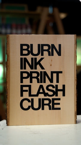 A piece of wood stands on a platen with words "Burn ink print flash cure" printed on it