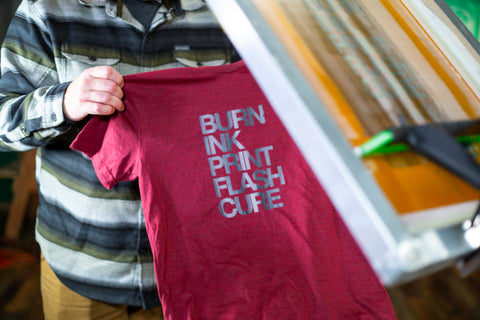 A man holds a red shirt next to a screen with black and white ink on it