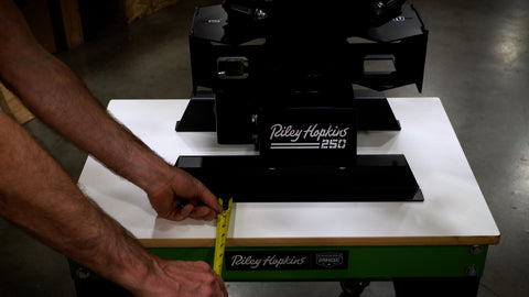 A riley hopkins 250 press sits on a cart. A man measures the distance from the cart to the end of the table