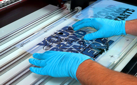 Dos manos con guantes de látex azul sostienen una película de impresión de pantalla sobre una mesa de luz en un cuarto oscuro